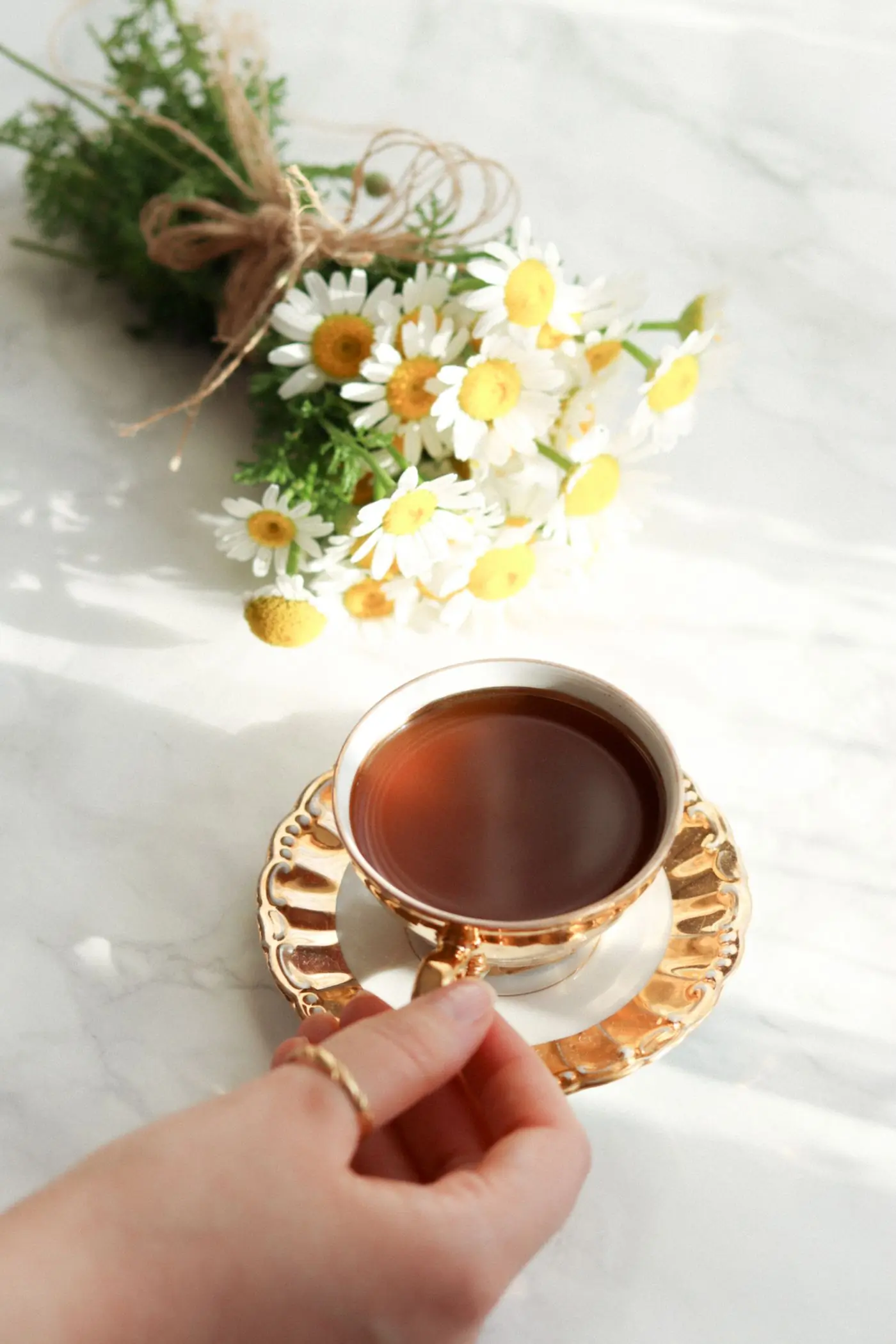 A hand lifting a cup of tea beside a small bunch of chamomile